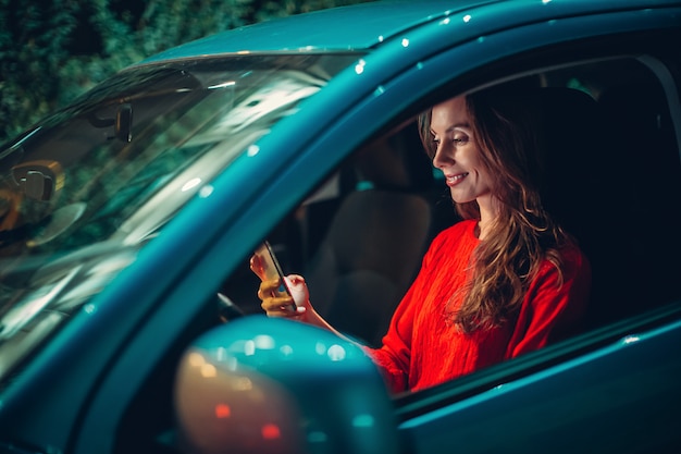 Feliz mulher europeia sentar no carro e usar smartphone