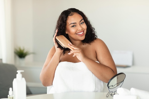 Feliz mulher gordinha negra escovando o cabelo encaracolado com pente e fazendo rotina de cuidados capilares em casa