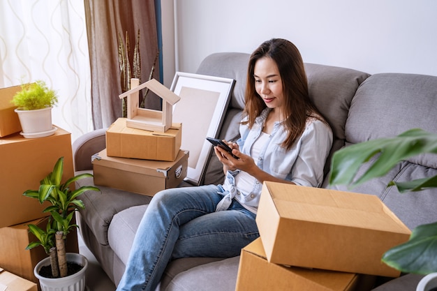 Feliz mulher jovem asiática usando smartphone na sala de estar em casa nova com pilha de caixas de papelão