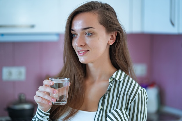 Feliz mulher jovem e atraente segurando um copo de água limpa e purificada na cozinha em casa