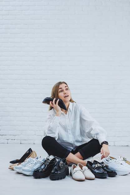 Feliz mulher jovem e bonita com um sorriso em uma roupa elegante com uma camisa branca e calça preta senta-se no chão entre os sapatos e escolhe um novo par de sapatos para si mesma