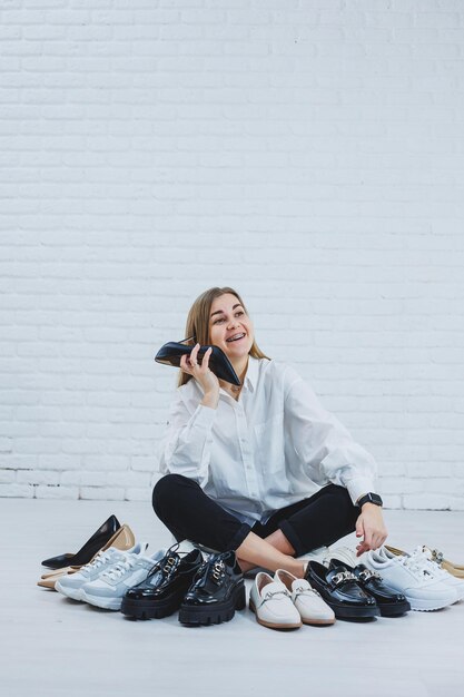 Feliz mulher jovem e bonita com um sorriso em uma roupa elegante com uma camisa branca e calça preta senta-se no chão entre os sapatos e escolhe um novo par de sapatos para si mesma