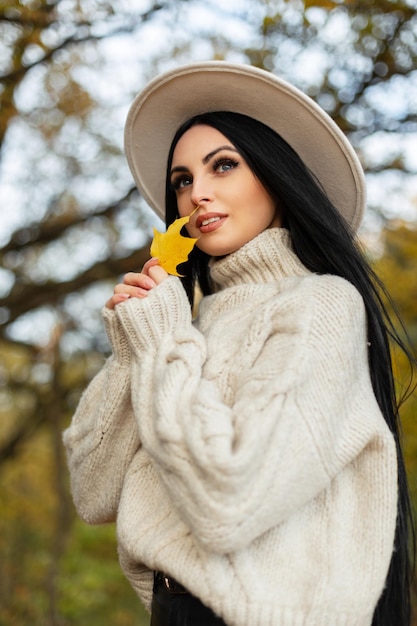 Feliz mulher jovem e bonita em uma roupa de malha vintage elegante com suéter bege e chapéu está segurando uma folha amarela de outono e andando no parque
