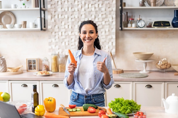 Feliz mulher latina segurando cenoura e gesticulando o polegar para cima aprovando receita enquanto cozinha na cozinha