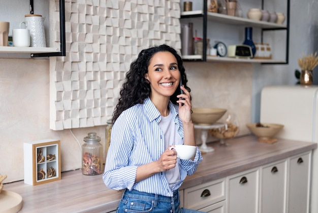 Feliz mulher latina tomando café e tendo telefonema em pé no interior da cozinha e sorrindo