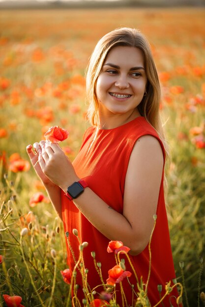 Feliz mulher loira com vestido vermelho, segurando uma flor no cabelo em um campo de papoulas. Vibrações divertidas de verão. Atmosfera descontraída, emoções positivas.