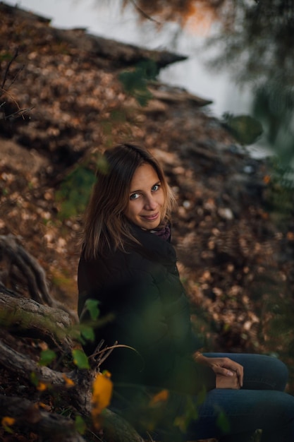 Feliz mulher loira sentada nas raízes das árvores na floresta sorrindo olhando para a câmera e posando perto do rio com árvore no fundo vestindo jeans azul e jaqueta preta caminhada de outono na floresta