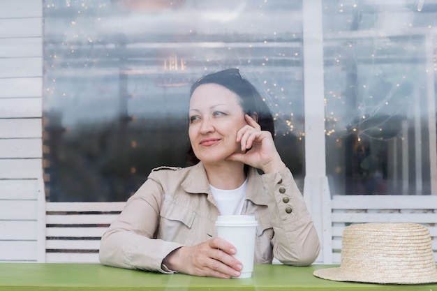 Feliz mulher madura com um copo de café se senta em uma mesa na varanda de verão de um café e sorrisos.