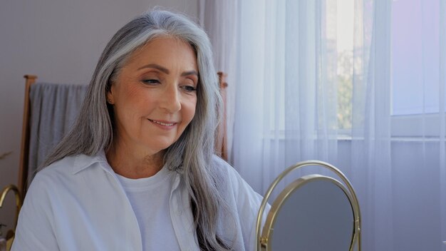 Feliz mulher madura de cabelos grisalhos olhando para o reflexo do espelho sorrindo satisfeita com a beleza natural