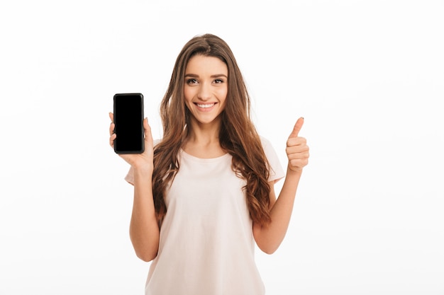 Feliz mulher morena de camiseta aparecendo a tela do smartphone em branco e o polegar enquanto sobre parede branca