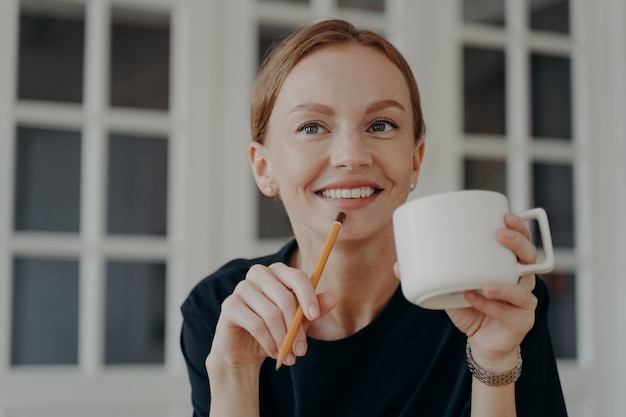 Feliz mulher pensativa segurando lápis bebe café da caneca sonhando pensando em conquistas de objetivos