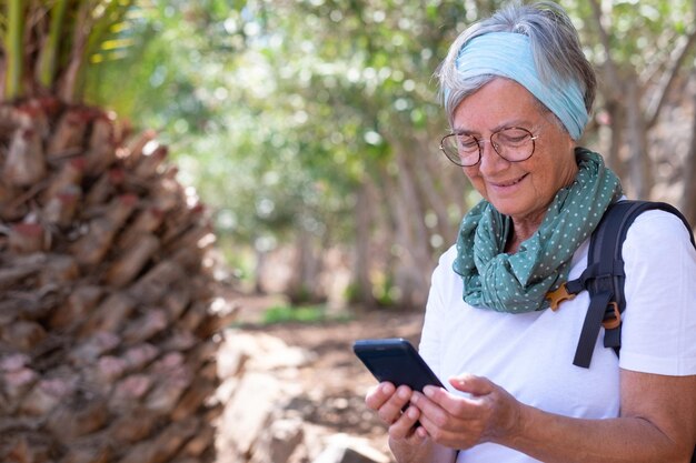 Feliz mulher sênior ativa com mochila em excursão ao ar livre sentado olhando para o celular Sorrisos femininos atraentes idosos desfrutando de estilo de vida saudável e dia ensolarado