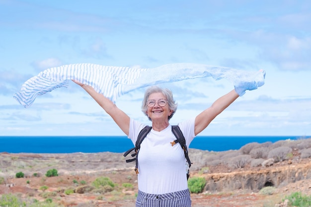 Feliz mulher sênior ativa com mochila enquanto caminhava ao ar livre acenando cachecol com braços estendidos Mar azul e céu ao fundo