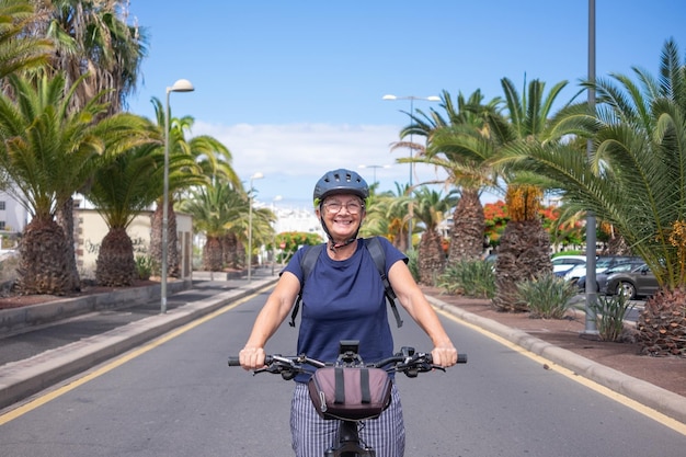 Feliz mulher sênior atraente com capacete e mochila desfrutando de atividade esportiva com bicicleta elétrica