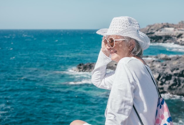 Feliz mulher sênior sorridente, de branco, usando chapéu e óculos de sol, sentada perto da praia, olhando para o horizonte sobre o mar, senhora idosa gosta de aposentadoria ou férias em dia ensolarado