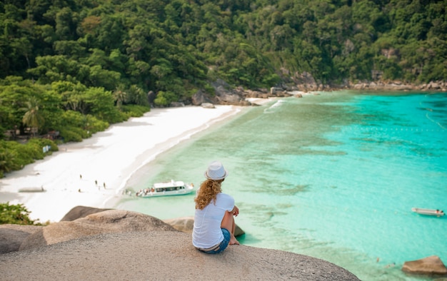 Feliz mulher sentada em uma pedra grande na praia tropical em camisa branca e chapéu de vime