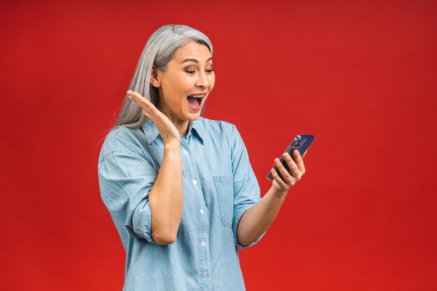 Feliz mulher vencedora asiática madura espantada e chocada segurando smartphone usando aplicativos móveis on-line sorrindo velha senhora de meia idade enviando mensagens de texto sms conversando no telefone isolado sobre fundo vermelho