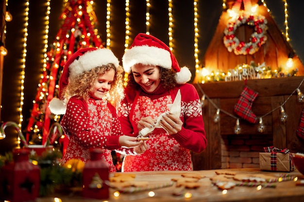 Feliz natal e boas festas. alegre encaracolada menina bonitinha e sua irmã mais velha em chapéus de papai noel cozinhar biscoitos de natal.