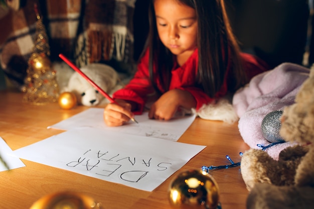 Feliz Natal e Boas Festas. Criança menina bonitinha escreve a carta para o Papai Noel perto da árvore de Natal em casa interior. O feriado, a infância, o inverno, o conceito de celebração