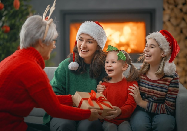 Feliz Natal e Boas Festas. Crianças alegres apresentando presentes para mamãe e vovó. Pais e filhos pequenos se divertindo perto de árvore dentro de casa. Família amorosa com presentes no quarto.