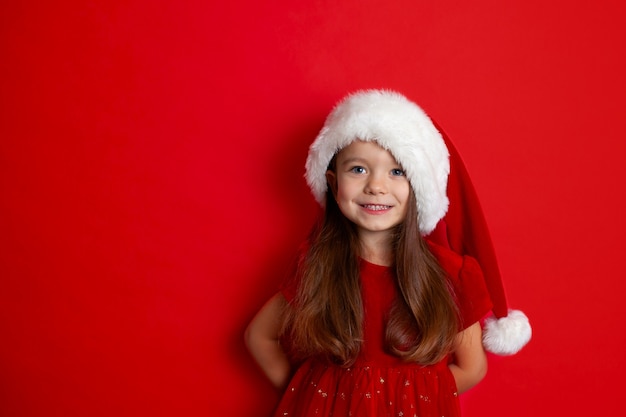 Feliz Natal e boas festas Retrato de uma menina em um boné de Papai Noel em um fundo vermelho Crianças