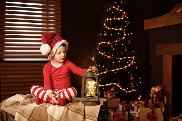 Foto feliz natal e boas festas um garotinho está sentado com uma lanterna na árvore de natal ano novo, elfo.