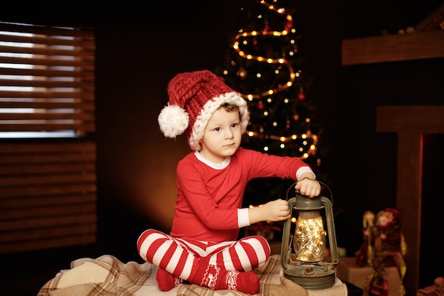 Foto feliz natal e boas festas um garotinho está sentado com uma lanterna na árvore de natal ano novo, elfo.