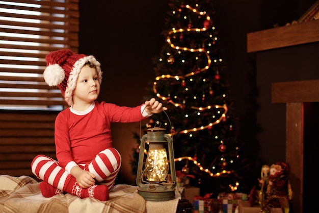 Foto feliz natal e boas festas um garotinho está sentado com uma lanterna na árvore de natal ano novo, elfo.