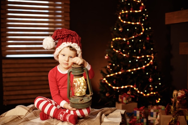 Foto feliz natal e boas festas um garotinho está sentado com uma lanterna na árvore de natal ano novo, elfo.