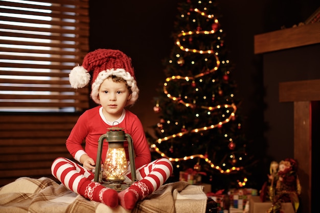 Foto feliz natal e boas festas um garotinho está sentado com uma lanterna na árvore de natal ano novo, elfo.