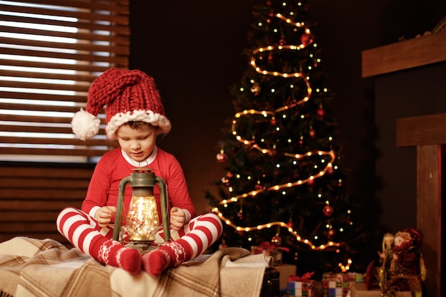 Feliz natal e boas festas um garotinho está sentado com uma lanterna na árvore de natal ano novo, elfo.