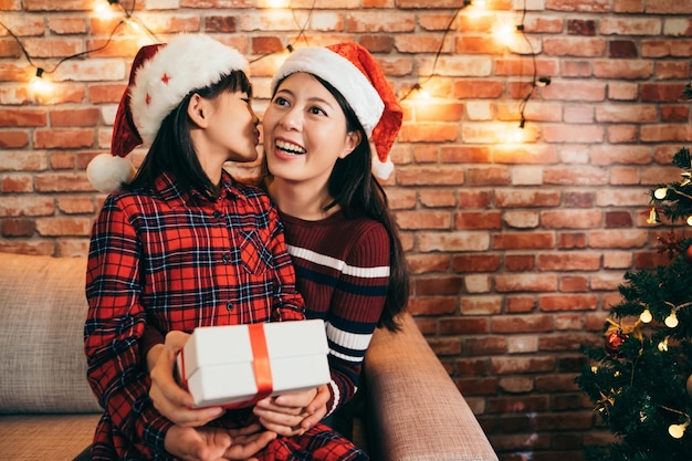 Feliz Natal e Boas Festas! uma mãe alegre surpreendendo seu filho com um presente de Natal. linda filha com chapéu de Papai Noel beijando a mãe em casa.