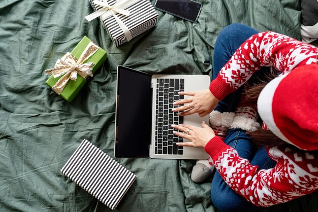 Feliz Natal e Feliz Ano Novo. Compras online. Mulher de vista superior vestindo roupas de Natal, sentada na cama usando o laptop. Tela de maquete, espaço de cópia