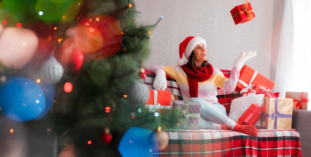Feliz Natal e Feliz Ano Novo Linda mulher morena com chapéu de Papai Noel e suéter está se divertindo com muitas caixas de presente no sofá em casa
