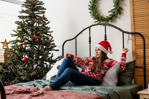 Feliz Natal e Feliz Ano Novo. Mulher jovem feliz com suéter vermelho e chapéu de Papai Noel sentada na cama comemorando o Natal