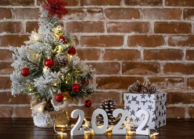 Feliz natal e feliz ano novo, pequena árvore de natal na mesa de madeira com bolas vermelhas e luzes. caixa de presente branca e 2022