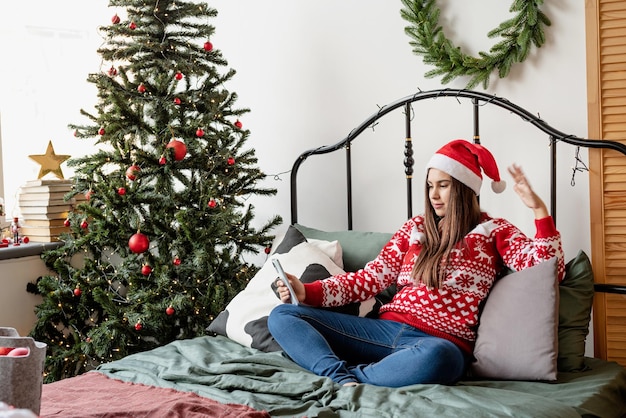 Feliz Natal e Feliz Ano Novo. Pessoas usando tecnologia. Jovem morena milenar de suéter vermelho e chapéu de Papai Noel, sentada na cama, usando mensagens de texto no celular ou fazendo uma videochamada