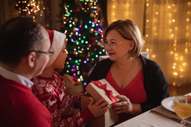 Feliz Natal! Família feliz comemorando o Natal em casa. Jantar festivo na árvore de Natal. Neto dando caixa de presente para a avó.