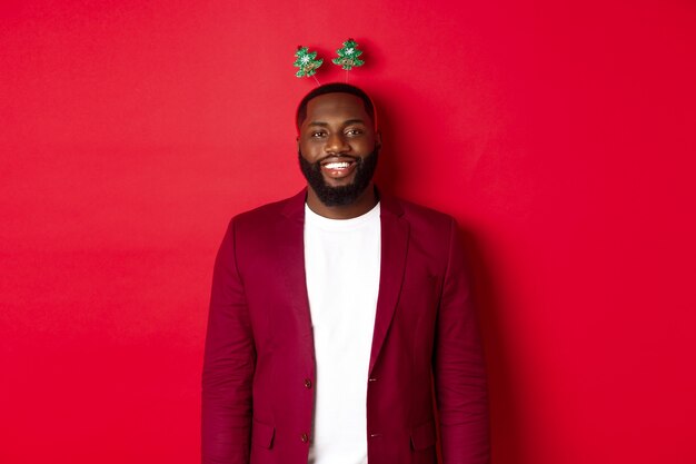 Feliz Natal. Feliz homem afro-americano celebrando o ano novo, usando uma bandana de festa engraçada e sorrindo, em pé sobre um fundo vermelho.