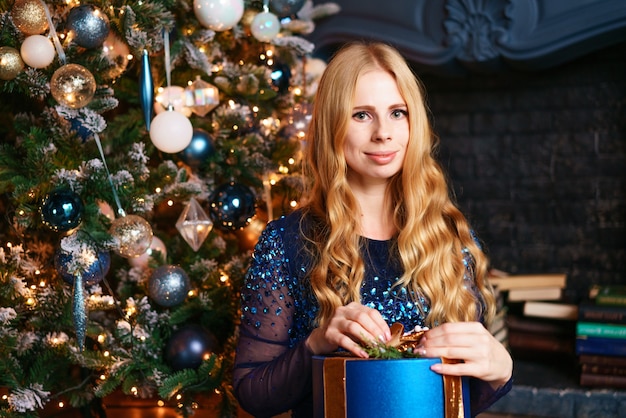 Feliz Natal linda garota caucasiana em um vestido azul incrível senta-se perto da árvore de Natal com dom segurar ...