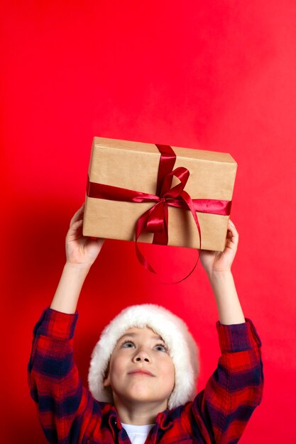 Foto feliz natal. menino de crianças segura uma caixa de presente amarrada com uma fita vermelha em um fundo vermelho. vertical. um lugar para texto. foto de alta qualidade