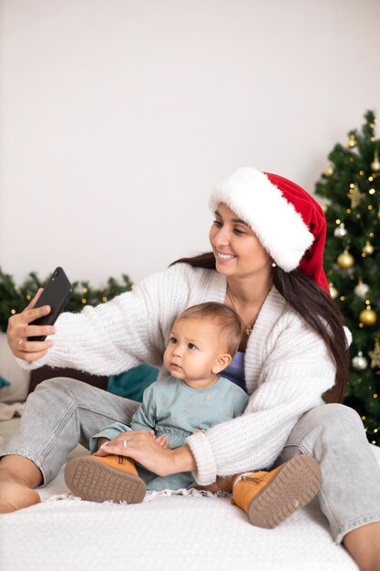 Feliz Natal Uma linda mãe e uma filhinha ligam para seus parentes via link de vídeo ou tiram uma selfie em um smartphone Quarto decorado aconchegante Estilo de vida