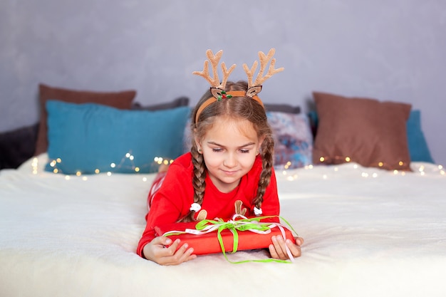 Feliz Navidad. Año nuevo. Feliz niña sonriente con coletas con caja de regalo de Navidad. niño en pijama rojo y con cuernos de venado en la cabeza se acuesta en la cama en Nochebuena y abre un regalo en casa.