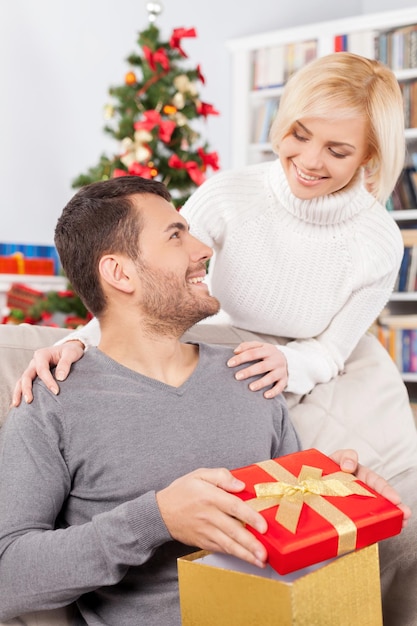 ¡Feliz Navidad! Apuesto joven sentado en el sofá y sosteniendo una caja de regalo mientras su novia está de pie detrás de él y sonriendo