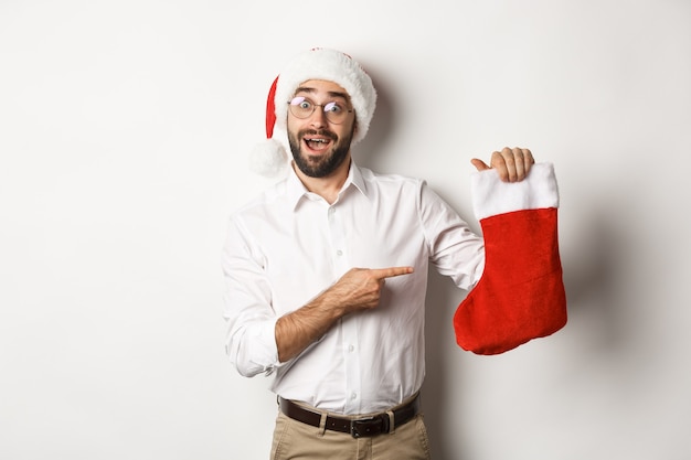 Feliz navidad, concepto de vacaciones. Hombre adulto feliz recibe regalos en calcetín de Navidad, mirando emocionado, con gorro de Papá Noel, fondo blanco.