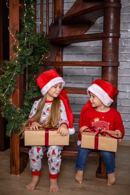 Feliz Navidad. Un dulce hermano y hermana con gorros de Papá Noel con cajas de regalo se sientan como en casa en las escaleras decoradas para Navidad. Foto de alta calidad