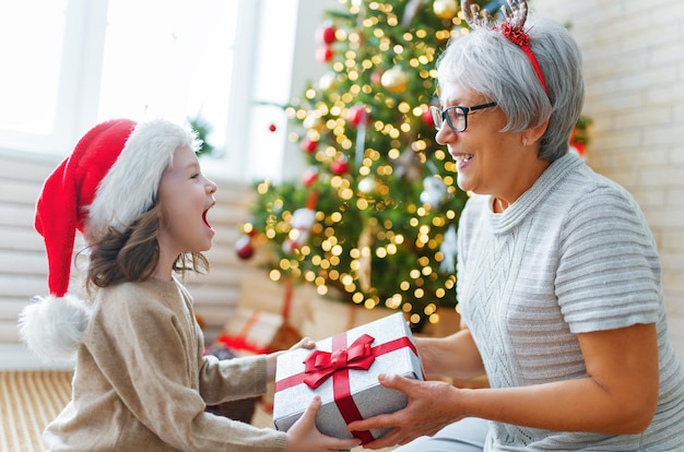 ¡Feliz Navidad y Felices Fiestas! Abuela alegre y su linda nieta intercambiando regalos. Abuelita y niño divirtiéndose cerca del árbol en el interior. Familia cariñosa con regalos en la habitación.