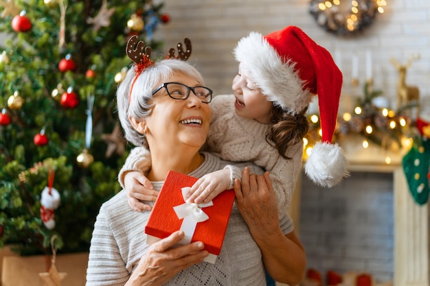 ¡Feliz Navidad y Felices Fiestas! Abuela alegre y su linda nieta intercambiando regalos. Abuelita y niño divirtiéndose cerca del árbol en el interior. Familia cariñosa con regalos en la habitación.