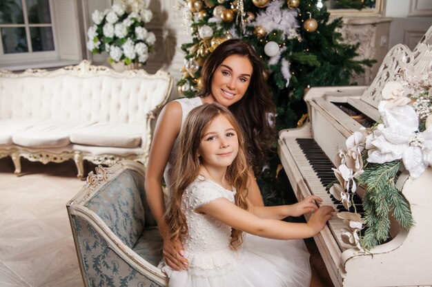 Feliz Navidad y felices fiestas. Alegre madre y su hija linda niña en interior clásico blanco jugando en un piano blanco decorado árbol de Navidad. Año nuevo