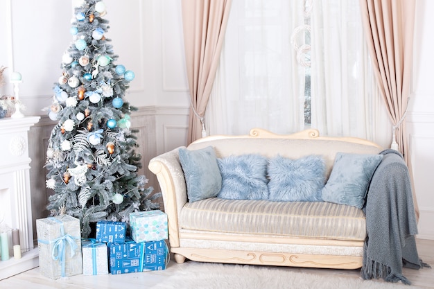 Feliz Navidad, felices fiestas. Elegante salón interior con árbol de Navidad decorado, chimenea y cómodo sofá. Árbol de Navidad con regalos a continuación. Interior de año nuevo.